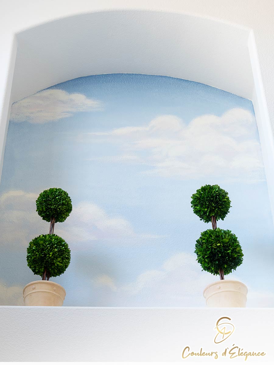 Two small topiaries sit in an archway with a painted cloud and sky background.